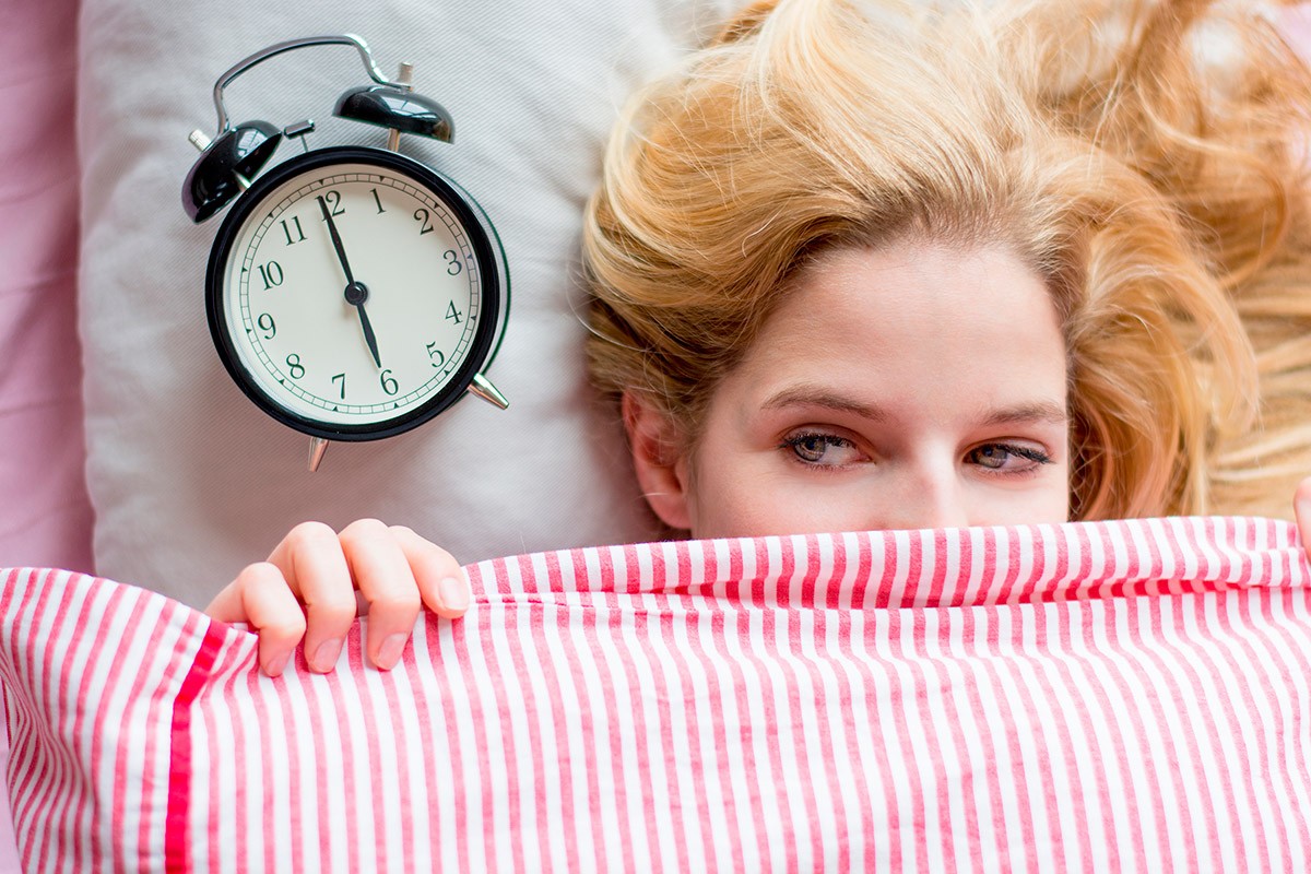 Femme blonde sous une couette avec un réveil sur la gauche de son oreiller